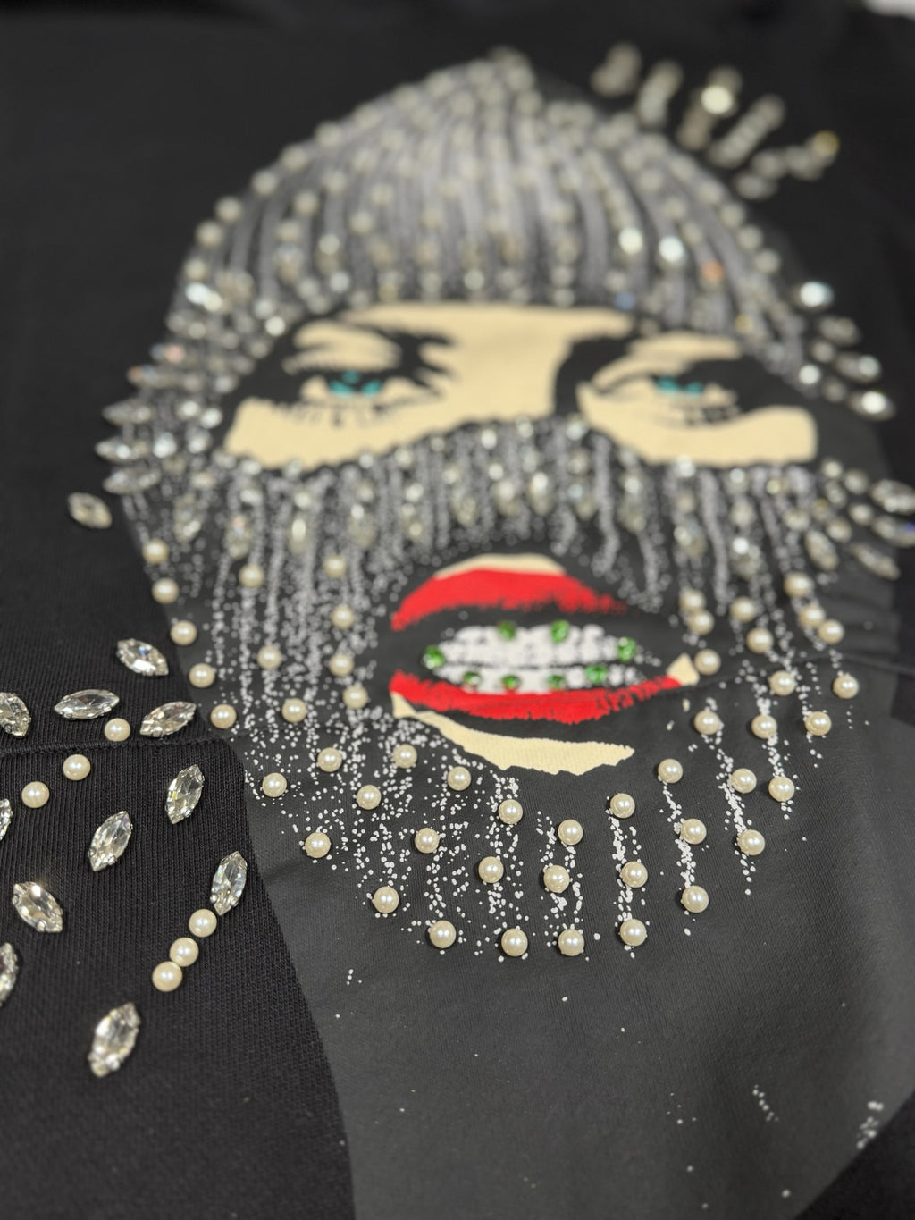 Dopetivity black hoodie featuring a graphic print of a masked face embellished with rhinestones and pearls.